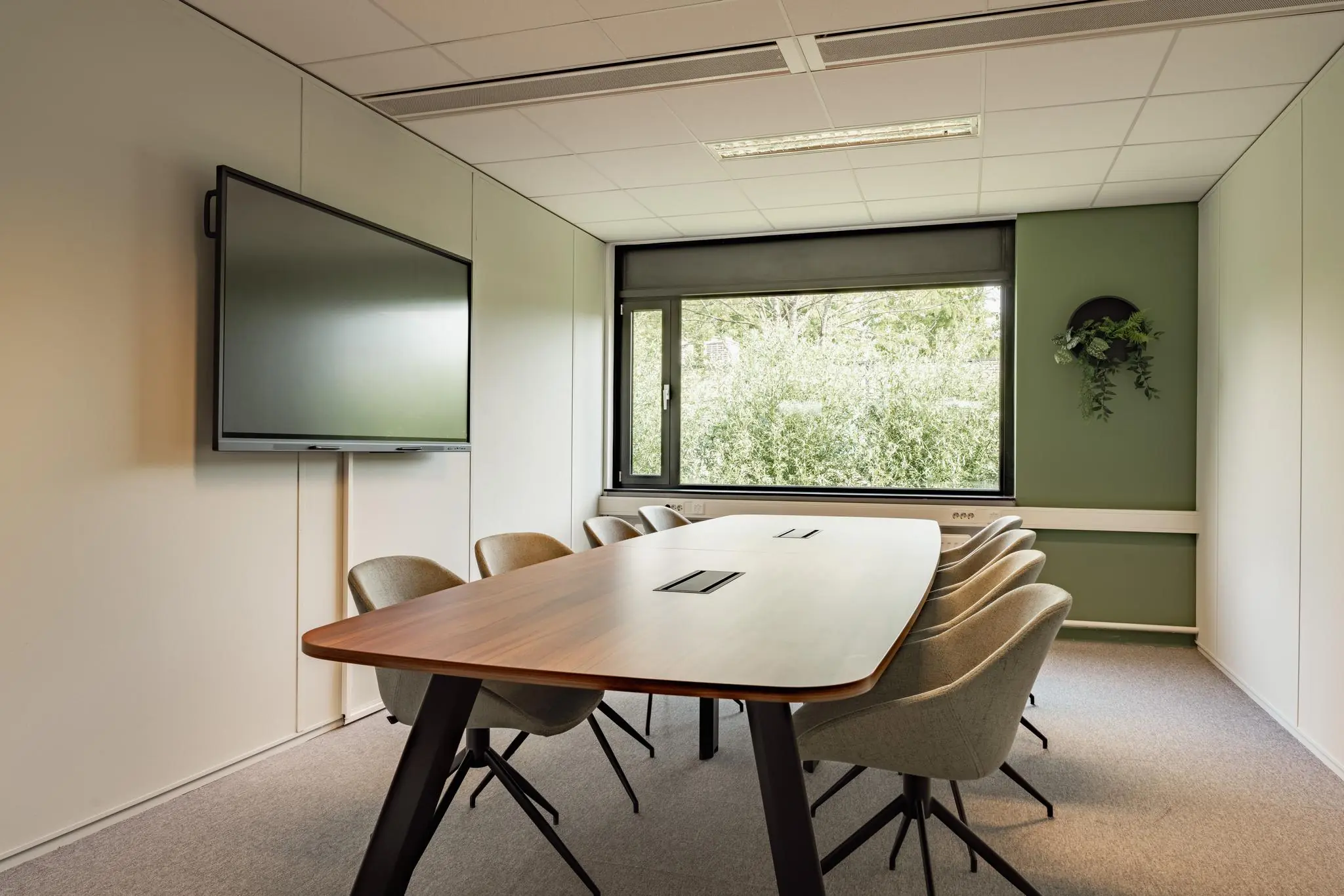 Vergaderruimte met een houten tafel, acht stoelen, een groot presentatiescherm en een raam met uitzicht op groen.